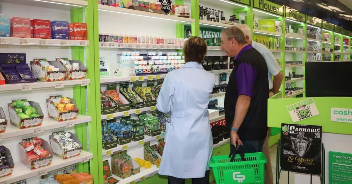 A store employee assists a customer browsing various cannabis products displayed on shelves in a brightly lit dispensary.